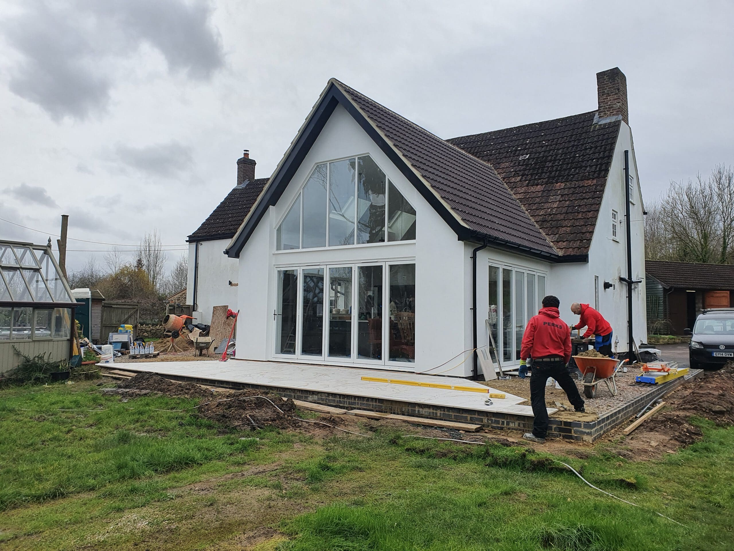 Two people work on building a patio outside a modern white house with large glass doors. Construction materials and tools are visible, and the yard appears muddy and grassy. The sky is overcast.