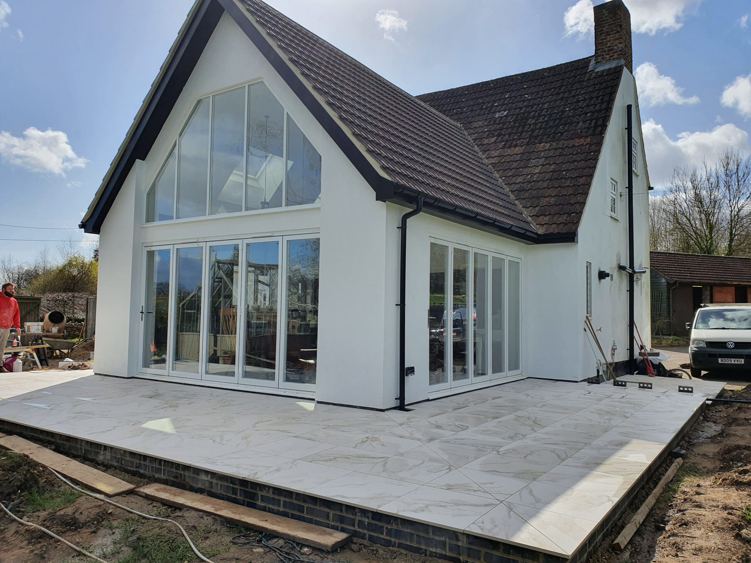 Modern white house with a steep, dark roof and large glass doors opening onto a spacious, newly tiled patio. The area is under construction, with tools and materials visible nearby.