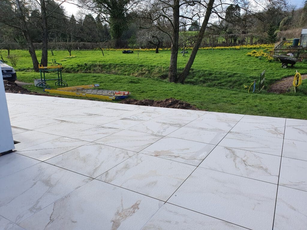 Newly installed large white patio tiles overlook a grassy yard with scattered trees, yellow flowers, garden tools, and equipment on a spring day. A trench runs parallel to the patio along the edge of the grass.