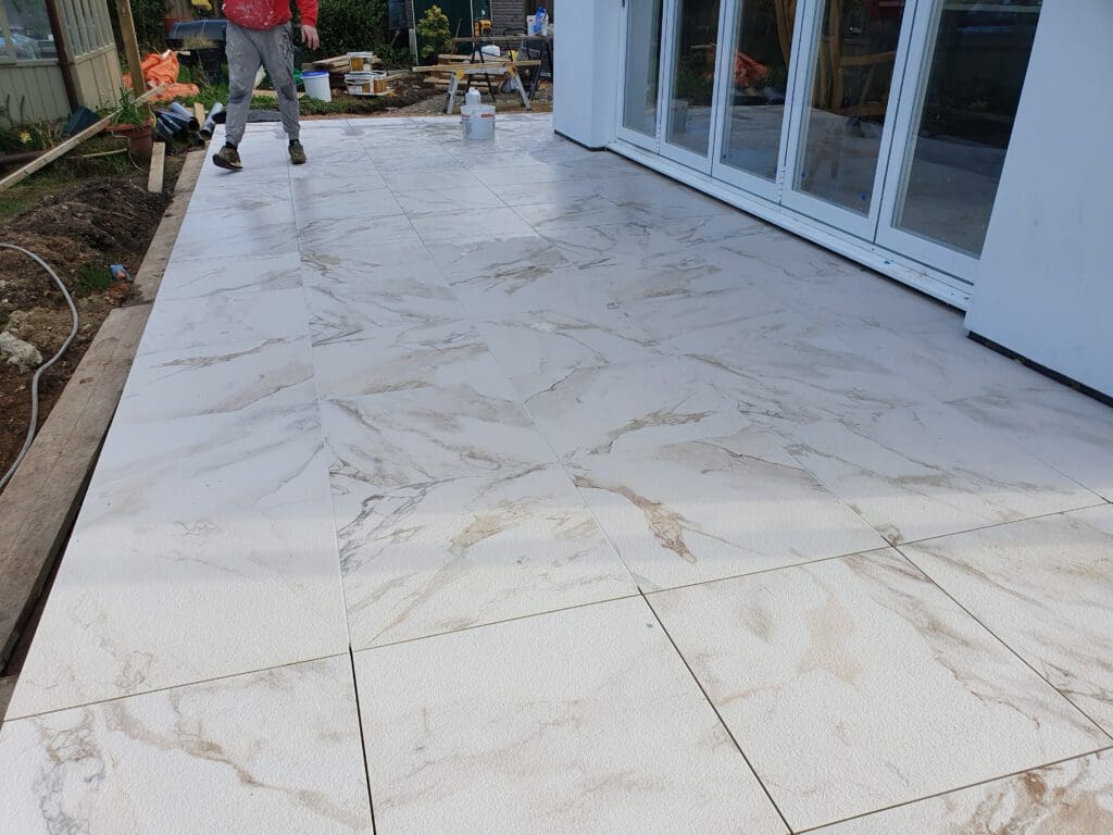 A newly tiled outdoor patio with large, light-colored tiles featuring a marble pattern. A person in work clothes stands nearby, and construction materials are visible in the background.