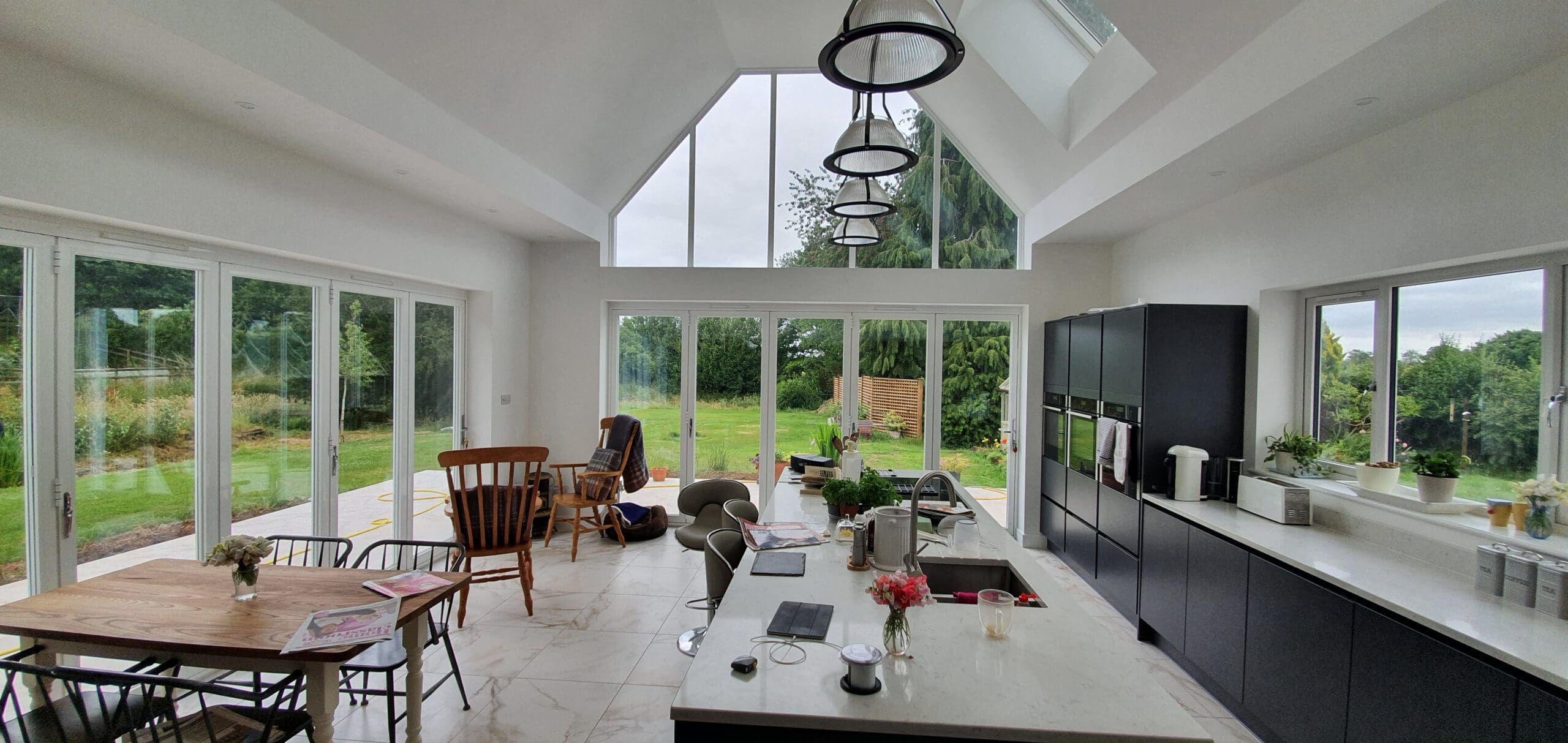 Modern, spacious kitchen with large windows and glass doors offering a view of a green garden. A wooden dining table with chairs, a kitchen island with flowers, and sleek black cabinets are visible under high ceilings.