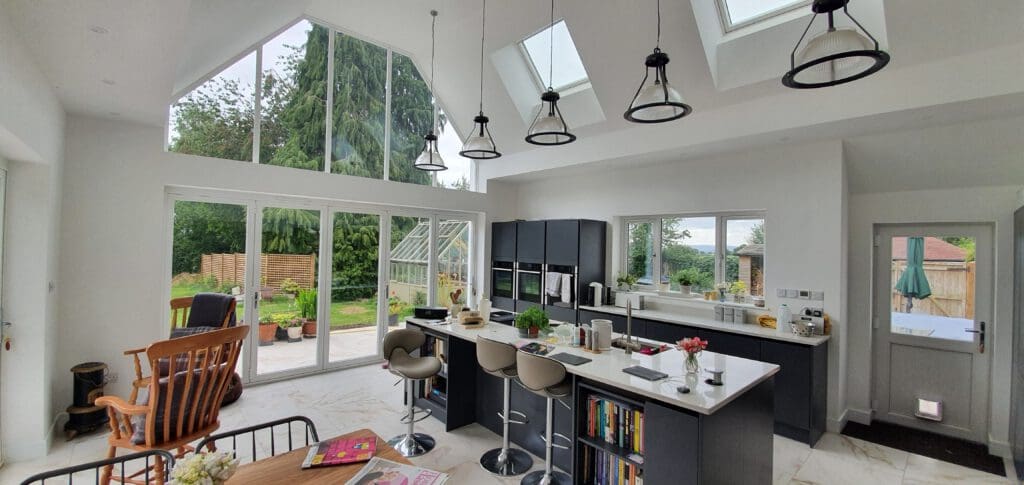 A modern, bright kitchen and living area with large windows and skylights, a central island with stools, dark cabinets, a breakfast bar, and views of a green garden outside.