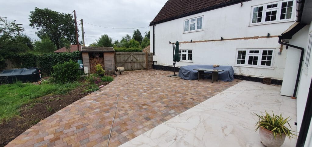Spacious backyard with a tiled patio, covered table and umbrella near a white house. There’s a grassy garden area, wooden shed, gate, and some potted plants. The sky is overcast.