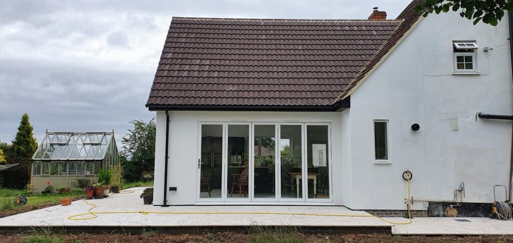 A white house with a brown tiled roof features large glass patio doors opening onto a new white-tiled patio. A yellow hose lies on the ground, and a greenhouse is visible in the garden to the left.