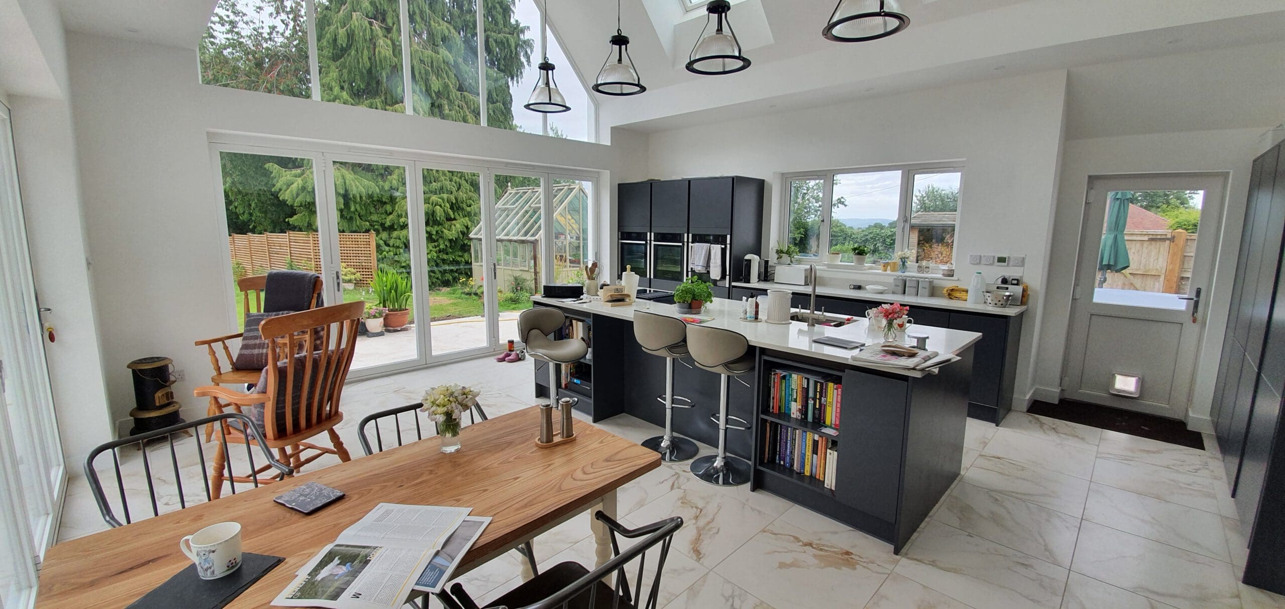 Modern kitchen with black cabinets, white countertops, and a central island with barstools. Large windows overlook a garden. A wooden dining table and rocking chair sit nearby on white tiled floors.