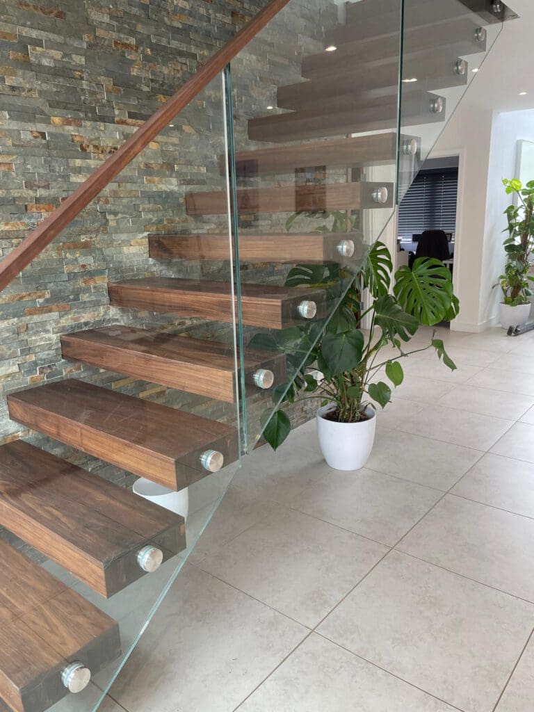 Modern floating wooden staircase with glass railing, next to a stone accent wall. There are potted green plants, including a monstera, on a light tiled floor in a bright, contemporary interior.