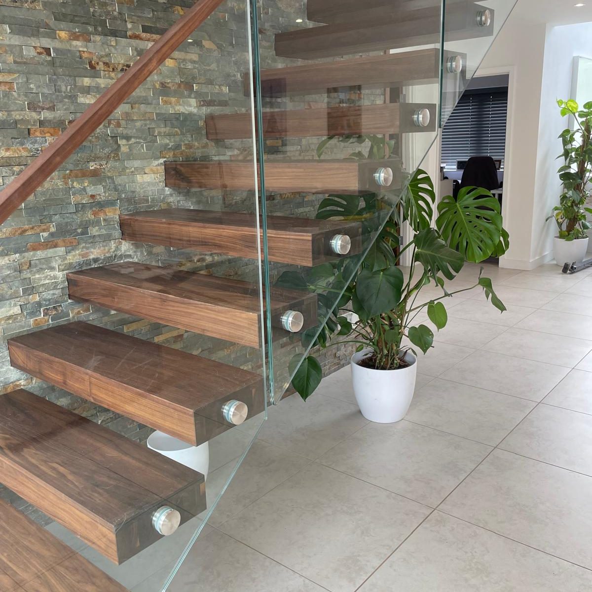 Modern floating wooden staircase with glass railing, next to a stone accent wall. There are potted green plants, including a monstera, on a light tiled floor in a bright, contemporary interior.