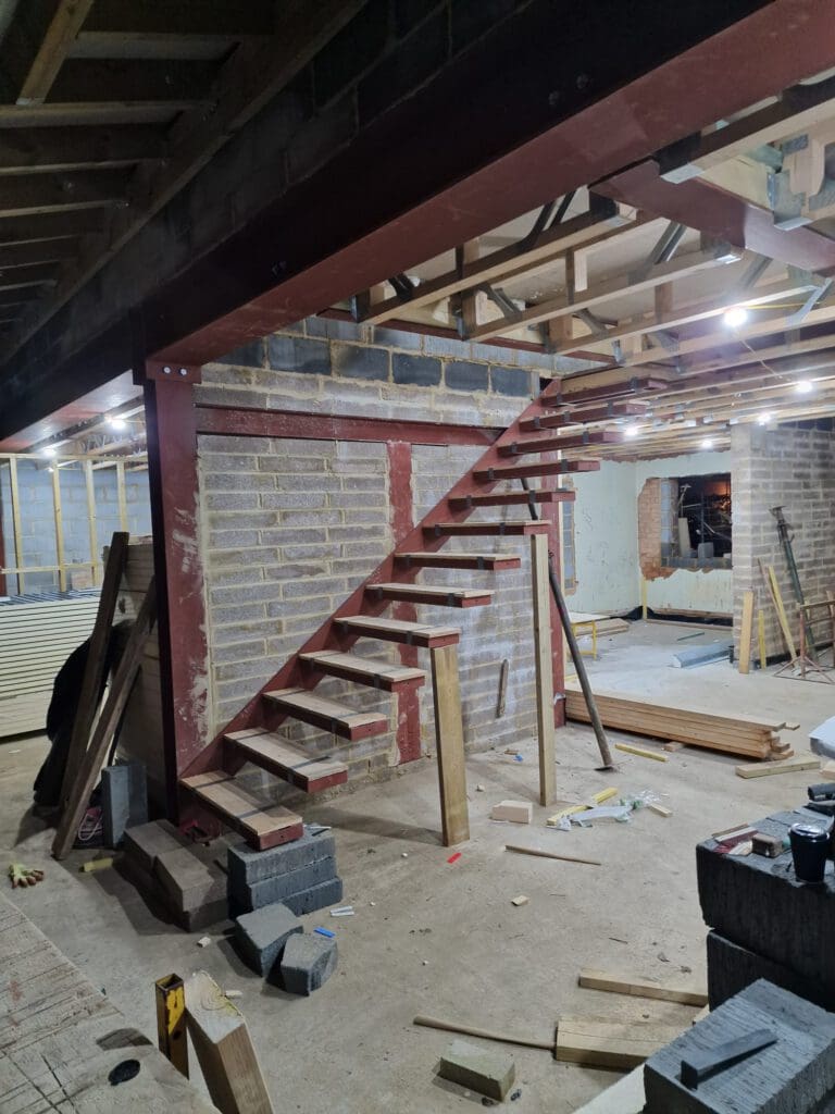 An unfinished staircase made of metal and wood beams is attached to a brick wall inside a construction site, with building materials and tools scattered on the floor. Ceiling lights illuminate the area.