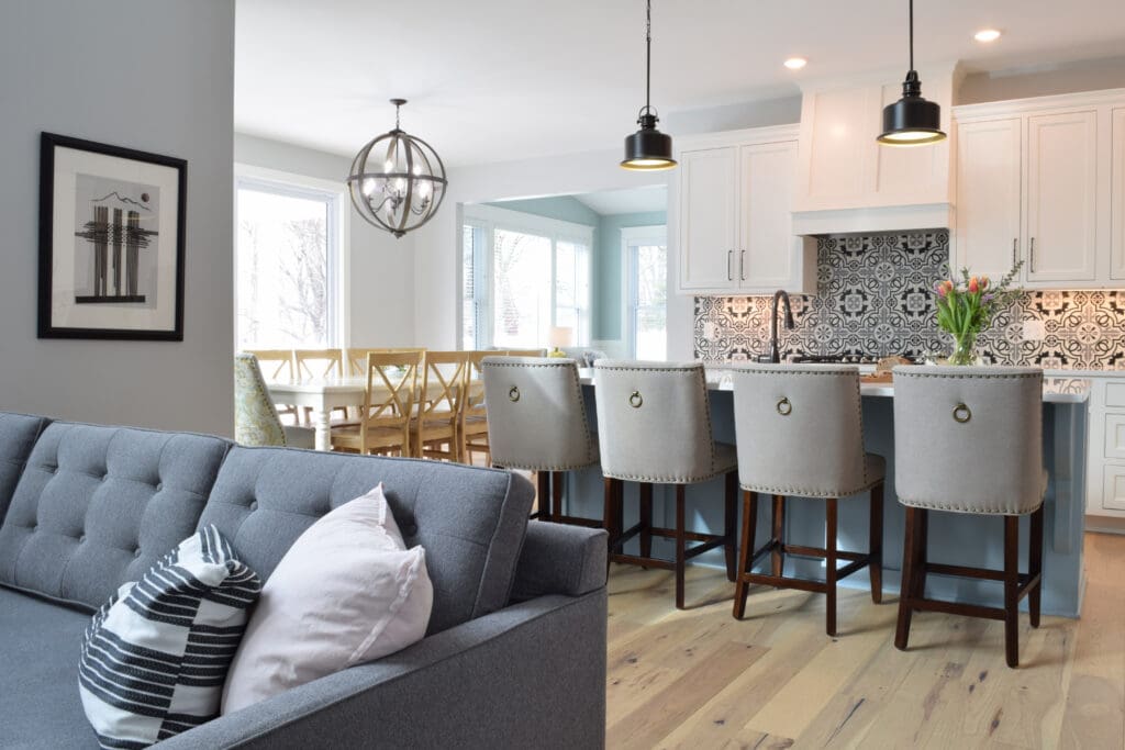 Modern open-concept kitchen and dining area with a gray sofa, patterned pillows, white cabinetry, black-and-white backsplash, upholstered bar stools, pendant lights, and a wooden dining table by large windows—recent structural renovations enhance the space.