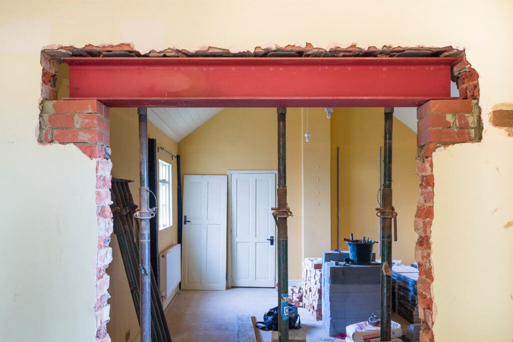 A partially demolished interior wall with exposed bricks and a red steel support beam installed above the opening, showcasing ongoing structural renovations; building materials and construction tools are visible in the background.