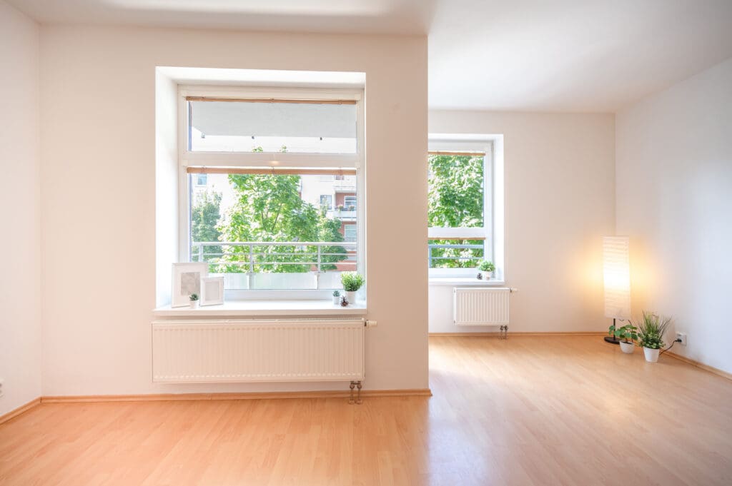 A bright, empty room with light wooden floors and large windows allows natural light to pour in, highlighting recent structural renovations. White walls and a few potted plants and decor items sit on the windowsill and floor.