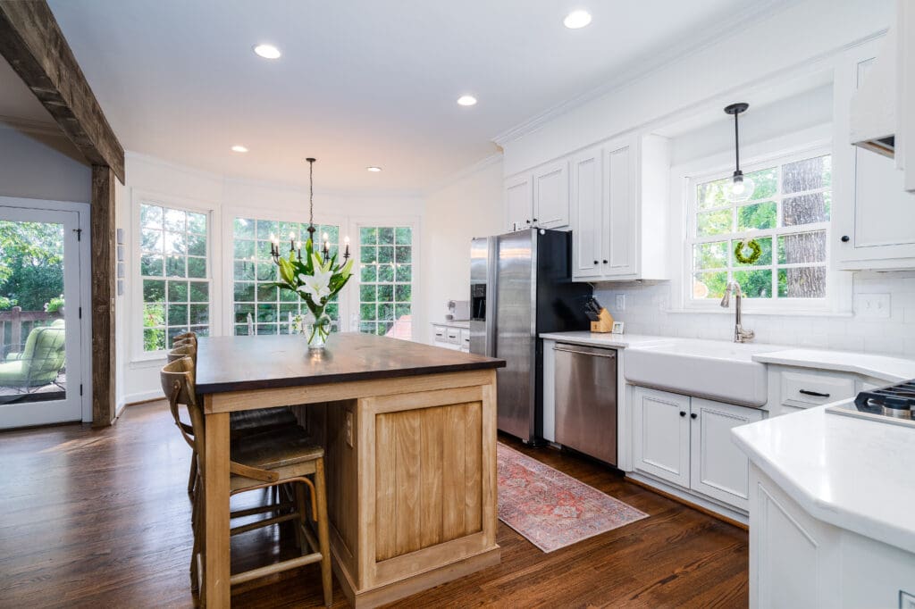 Bright, modern kitchen extension with large windows, white cabinets, stainless steel appliances, a wooden island with chairs, a farmhouse sink, hardwood floors, and a vase of flowers on the island.