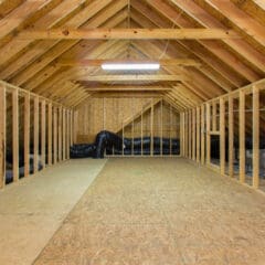 Unfinished attic with exposed wooden beams, plywood floor, and framed walls; ideal for structural renovations. Black HVAC ductwork runs along the walls, and a single fluorescent light fixture hangs from the ceiling.