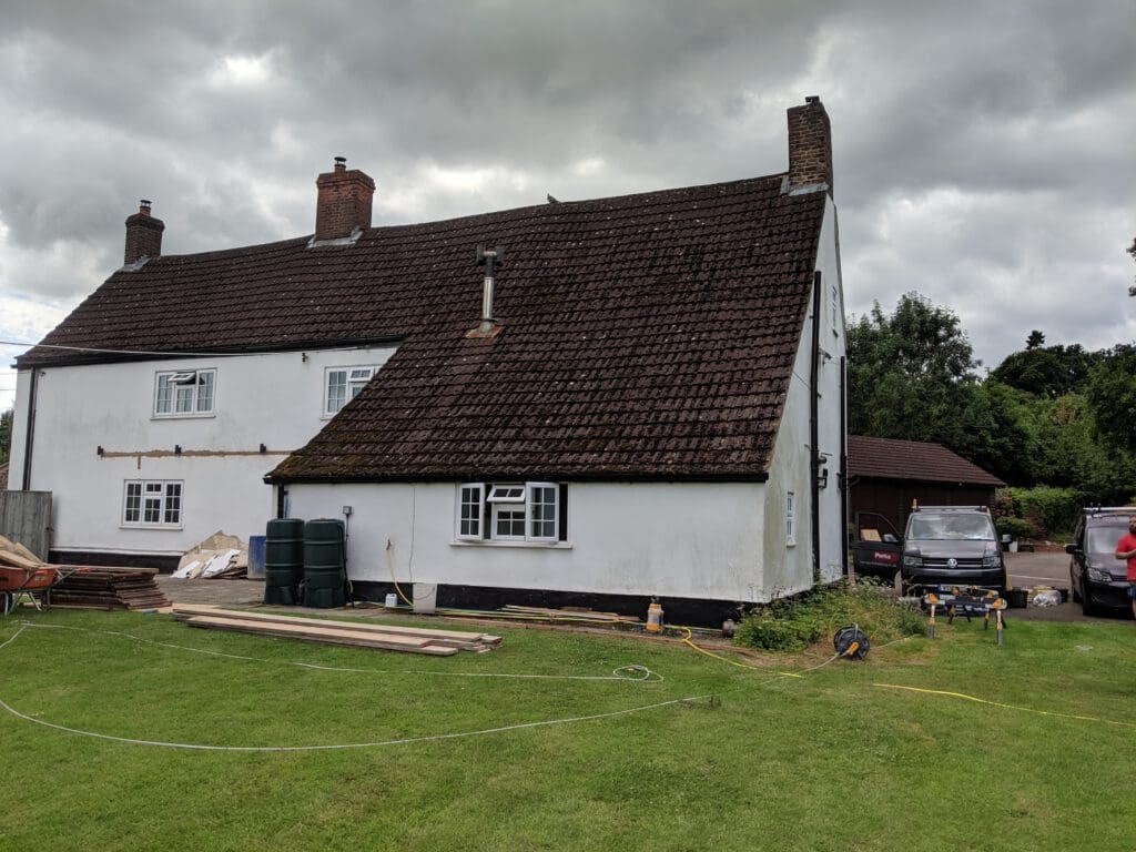 A white two-story house with a sloped brown tile roof, chimneys, and several small windows sits on a green lawn under a cloudy sky. Construction materials and a couple of vehicles are visible nearby.