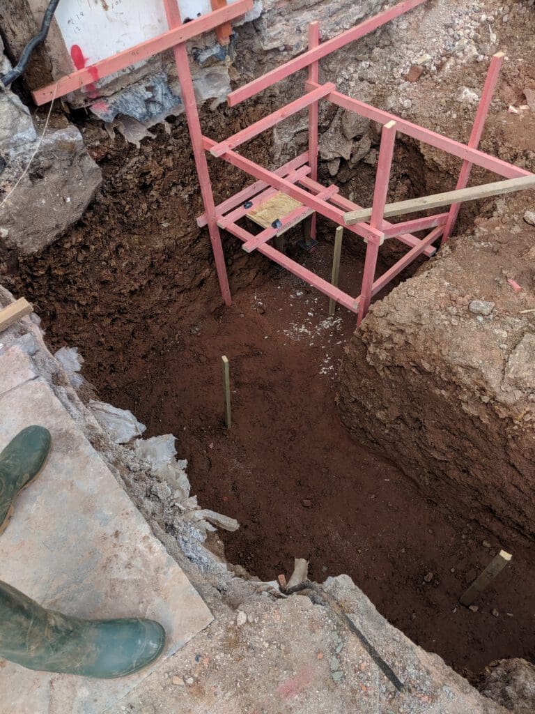 A deep construction pit with exposed soil and rocks, wooden stakes in the ground, and a temporary wooden framework. A person in green boots stands on the edge of the excavation.