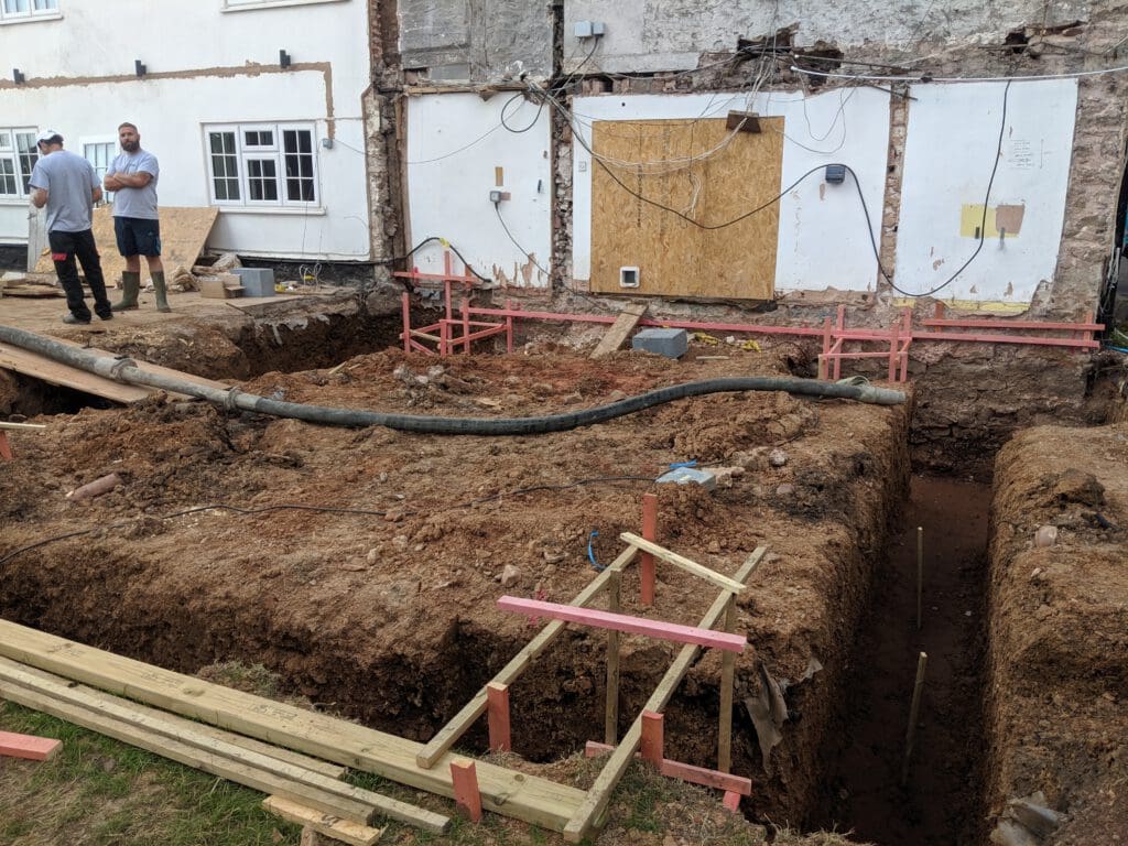 Construction site showing deep foundation trenches next to a building with exposed walls and wiring; two people stand on the left, while wooden and metal supports surround the excavated area.