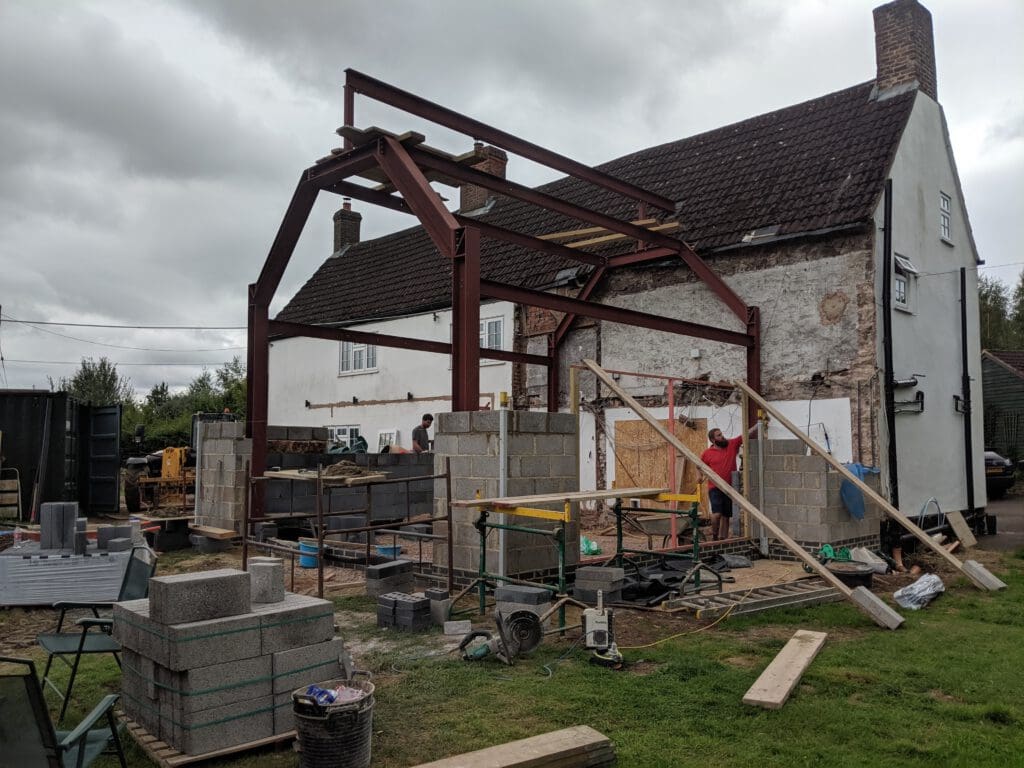 A house extension under construction with steel beams, cinder block walls, scaffolding, and building materials. Two people are working on the site beside the existing white house on a cloudy day.
