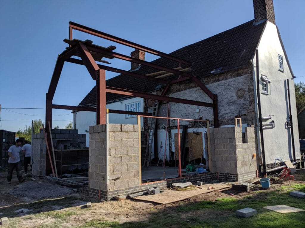 A partially constructed extension with steel beams and concrete block walls is attached to an old house. Two people stand nearby, and building materials are scattered around the site.