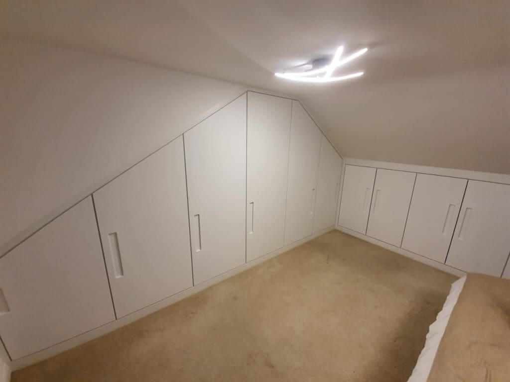 A room with beige carpet, slanted ceiling, and built-in white cabinets with vertical handles along the wall and under the eaves; a modern ceiling light is on.