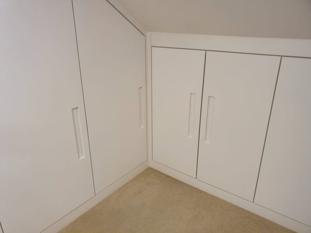 Built-in white cupboards with sleek handles are fitted into the corner of a room with a sloped ceiling. The cupboards are set against cream-colored carpet flooring.