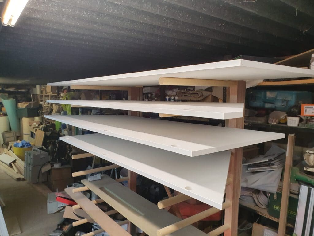 Several large white wooden boards are stacked horizontally on a makeshift rack in a cluttered workshop or garage, surrounded by various tools, materials, and storage shelves.