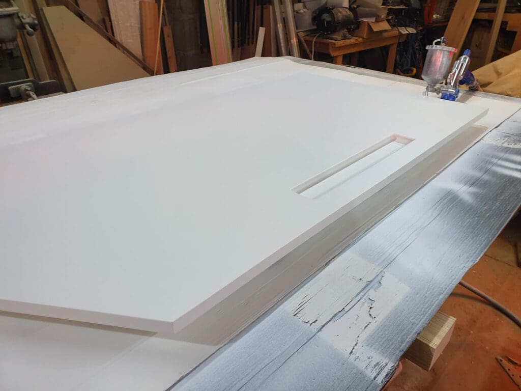 A white cabinet door with a rectangular handle cutout lies on a workbench in a woodworking shop, surrounded by tools and pieces of wood.