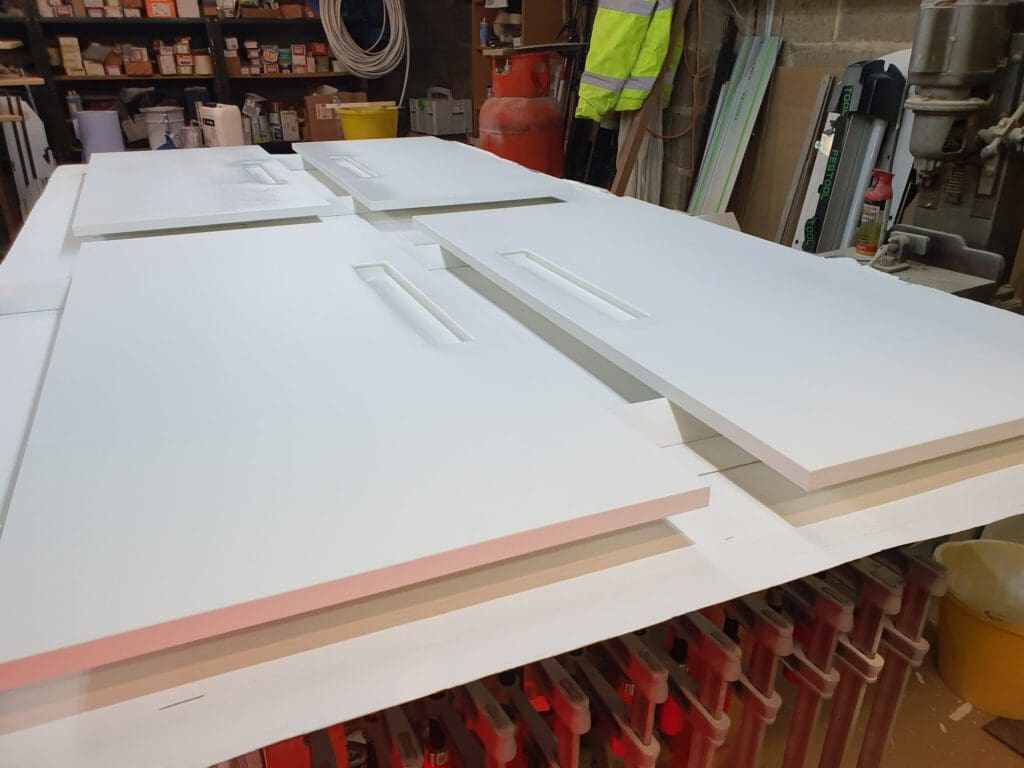 Several white cabinet doors with inset handles are laid flat on a workbench in a workshop, surrounded by tools, paint cans, and various supplies.