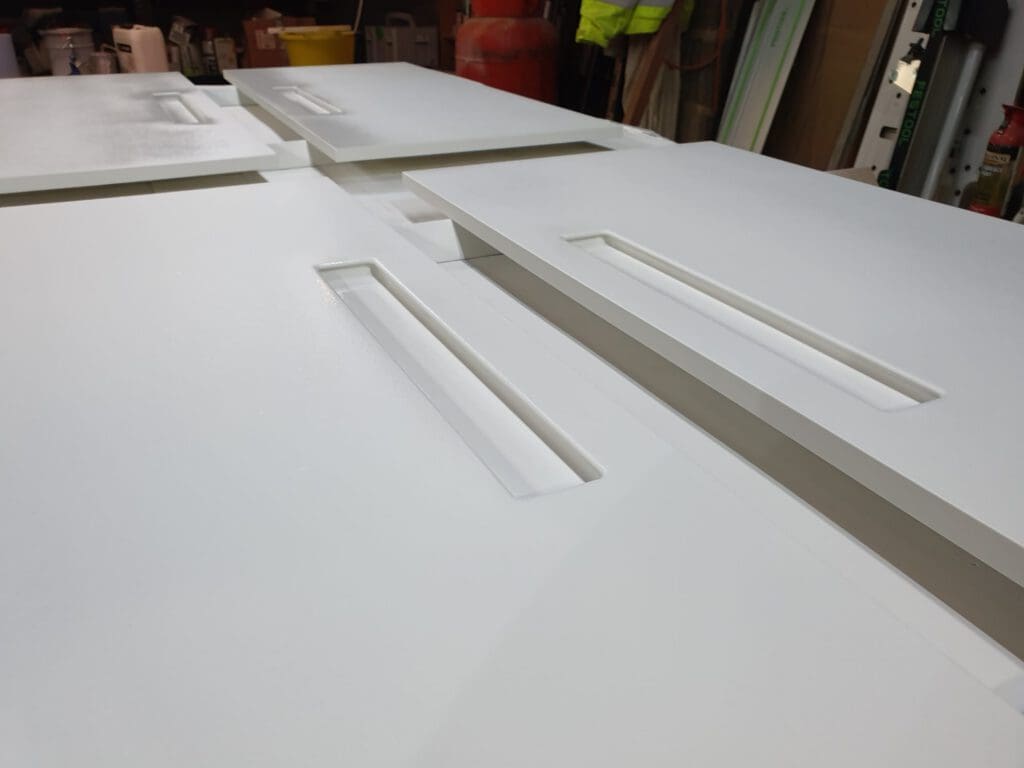 Several smooth, white cabinet doors with rectangular cutout handles are laid out flat, overlapping each other in a workshop setting with various tools and materials in the background.