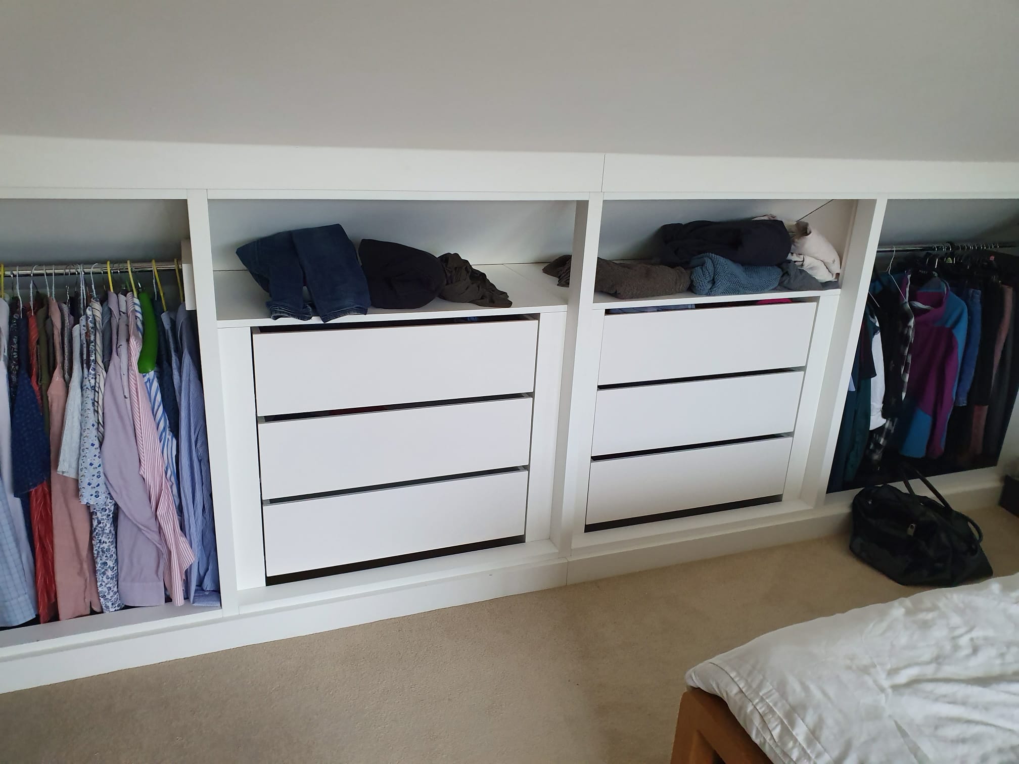 A built-in white wardrobe with two sets of drawers, shelves holding folded clothes, and hanging clothes on rails. Some shirts, jackets, and garments are neatly organized. The wardrobe is in a carpeted bedroom.