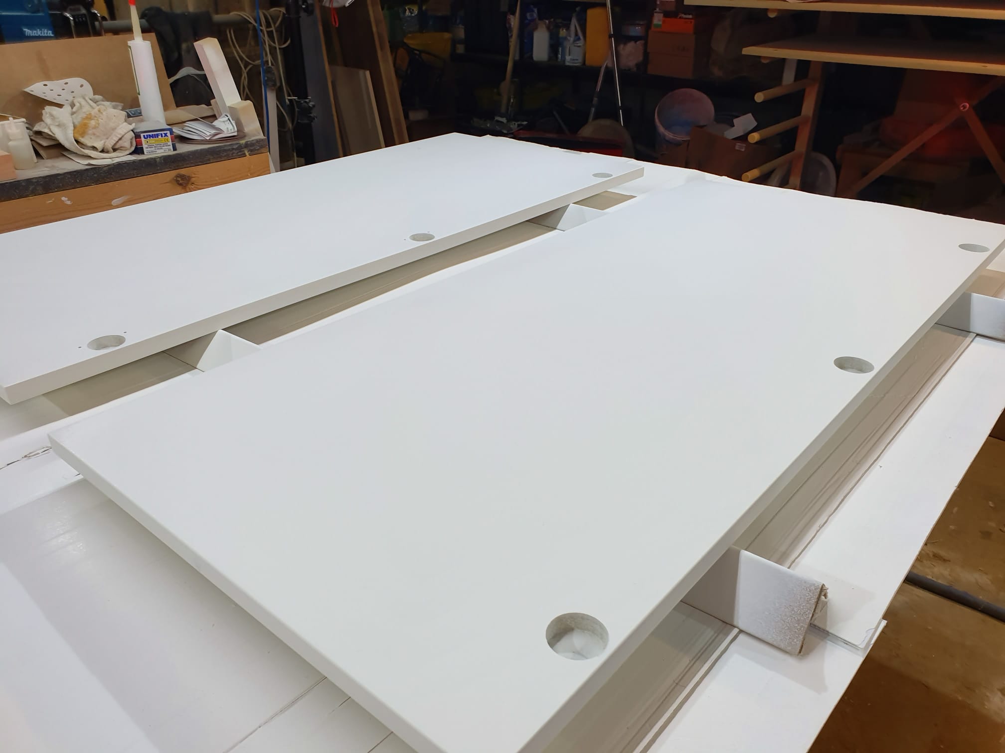Two large white rectangular boards with round holes near the edges rest on a workbench in a workshop filled with various tools and materials.