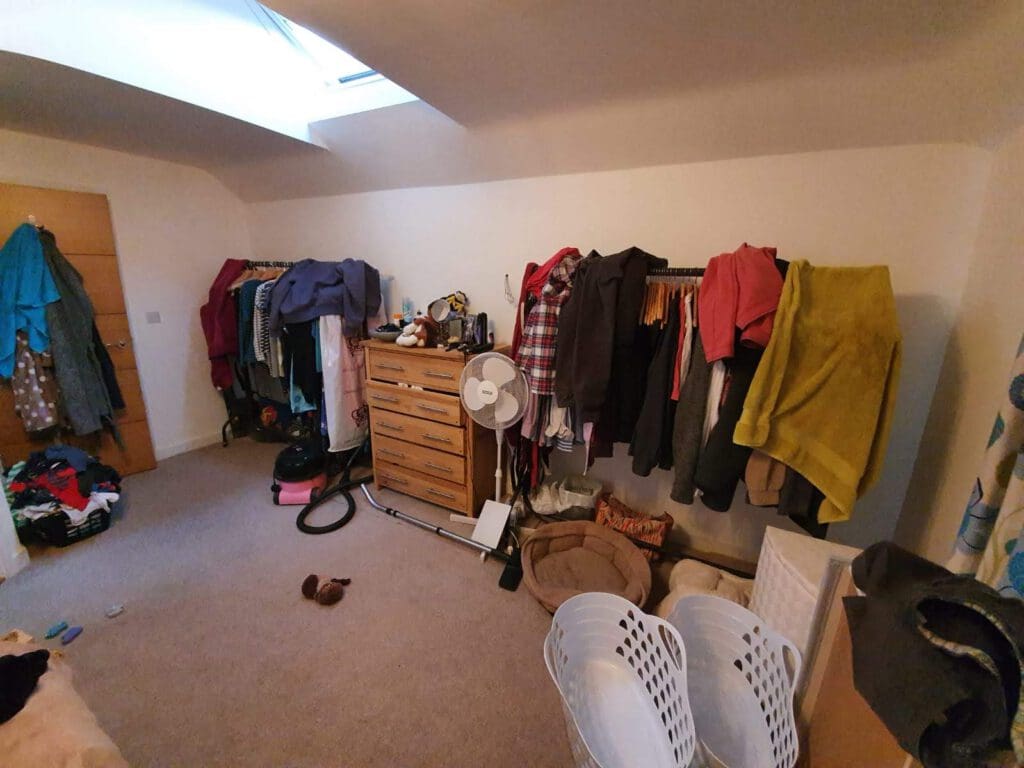 A cluttered bedroom with clothes hanging on racks, a wooden dresser, a fan, laundry baskets, and various items scattered around the floor, including a vacuum cleaner, shoes, and pet beds.
