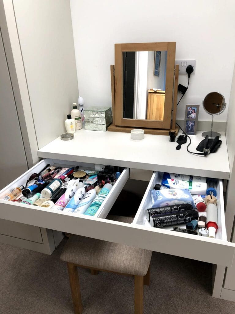 A white vanity table with an open drawer full of beauty and hair products, a wooden framed mirror, a stool, a hairdryer, brushes, bottles, and framed photos on top. The surrounding room is carpeted.