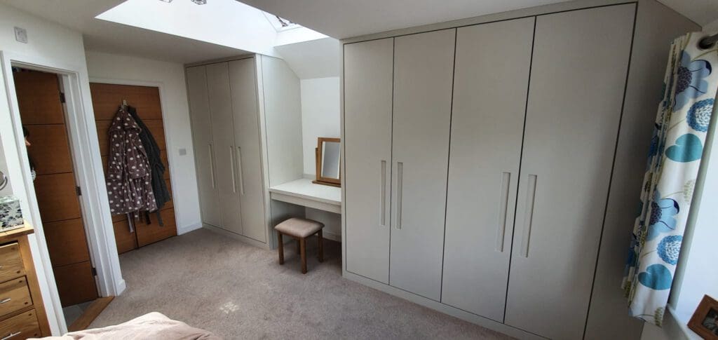Bedroom with beige fitted wardrobes, a small dressing table with a mirror and stool, a wooden door with coats hanging on hooks, light carpet flooring, and a window with patterned curtains.