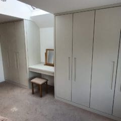 Bedroom with beige fitted wardrobes, a small dressing table with a mirror and stool, a wooden door with coats hanging on hooks, light carpet flooring, and a window with patterned curtains.