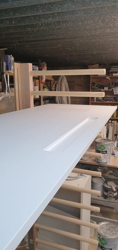 A white door with a rectangular cut-out handle is propped up in a workshop. Shelves with paint cans, tools, and other supplies are visible in the background.