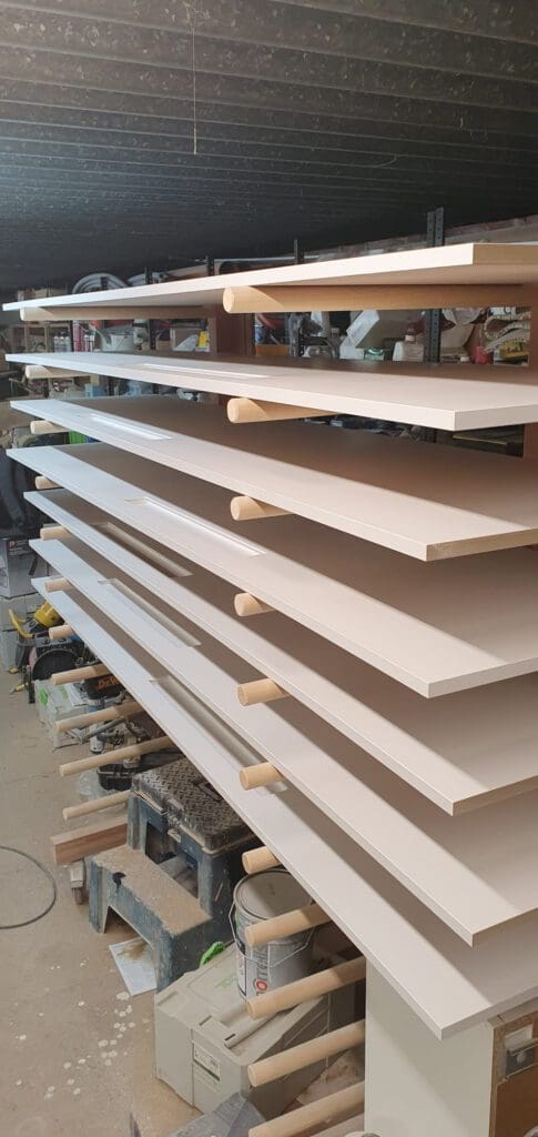 Several large, flat wooden boards are stacked horizontally with gaps between them, supported by round dowels, in a workshop filled with tools, wood, and construction materials.