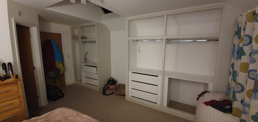 A bedroom with built-in open wardrobes, drawers, and a clothes rail. There are towels hanging on the door, a few items on the floor including a vacuum, a chest of drawers, and patterned curtains by a window.