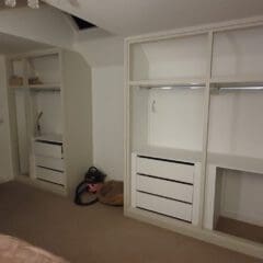 A bedroom with built-in open wardrobes, drawers, and a clothes rail. There are towels hanging on the door, a few items on the floor including a vacuum, a chest of drawers, and patterned curtains by a window.