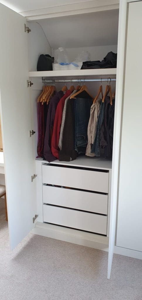An open white wardrobe shows coats and shirts hanging on wooden hangers above three closed drawers, with bags and folded items stored on the top shelf.