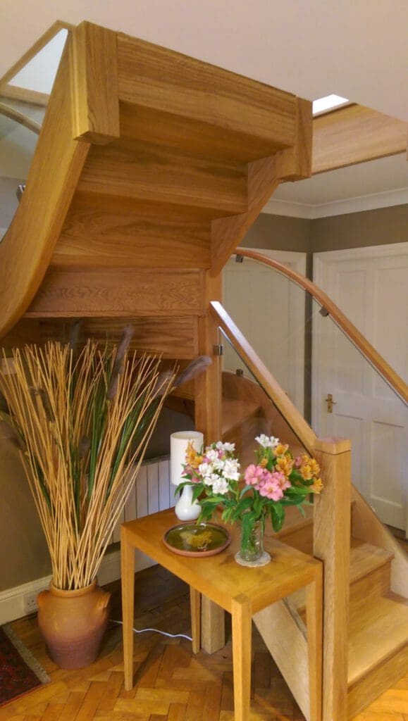 A wooden staircase with glass railing curves upward in a modern home. Below it, a table holds a vase of colorful flowers, a bowl, a small lamp, and a large pot with tall dried plants beside it.