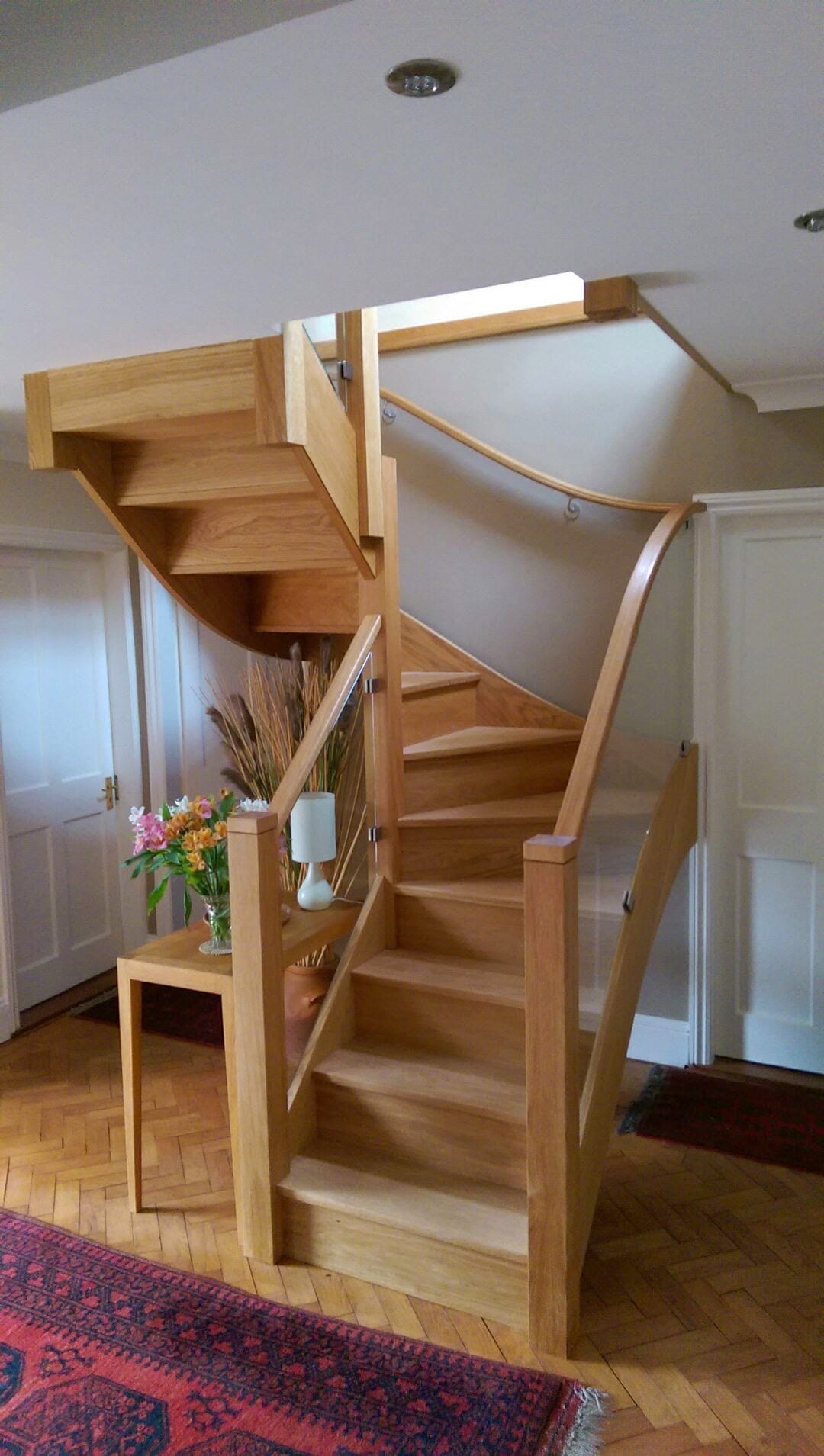 A modern wooden spiral staircase with glass railing curves upward in a bright hallway, next to a small table with a lamp and dried plants on a parquet floor with patterned rugs.