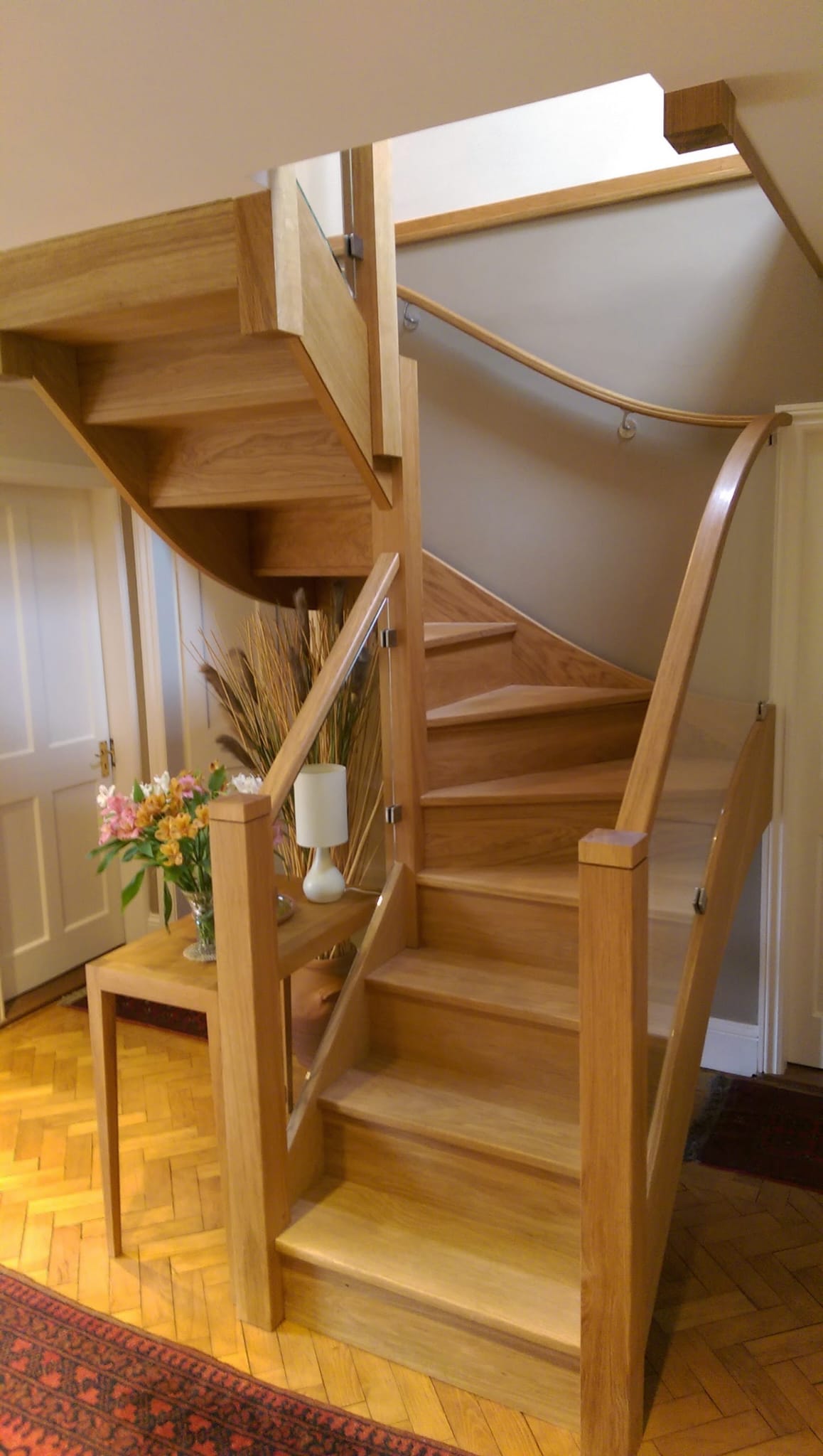A modern wooden staircase with a curved design leads upstairs. A small table with a lamp, vase of flowers, and decorative plants sits to the left on a herringbone wood floor. A red rug is partially visible.