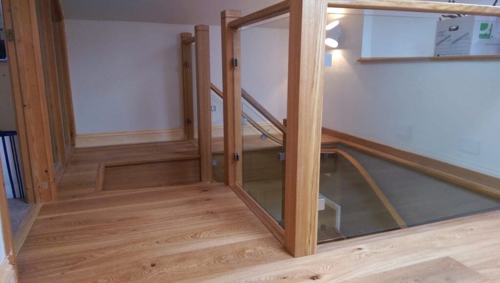 A modern wooden loft floor with glass-paneled railings, leading to a staircase going down. The space is well-lit with light wood tones and minimalist decor.