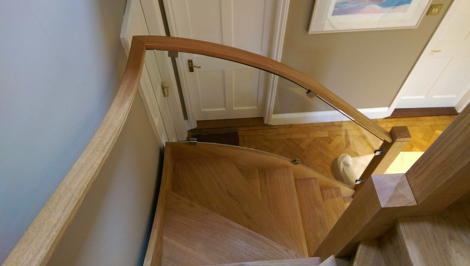 A wooden spiral staircase with a curved handrail and glass panel, leading down to a hallway with parquet flooring and white doors. A framed picture hangs on the beige wall.
