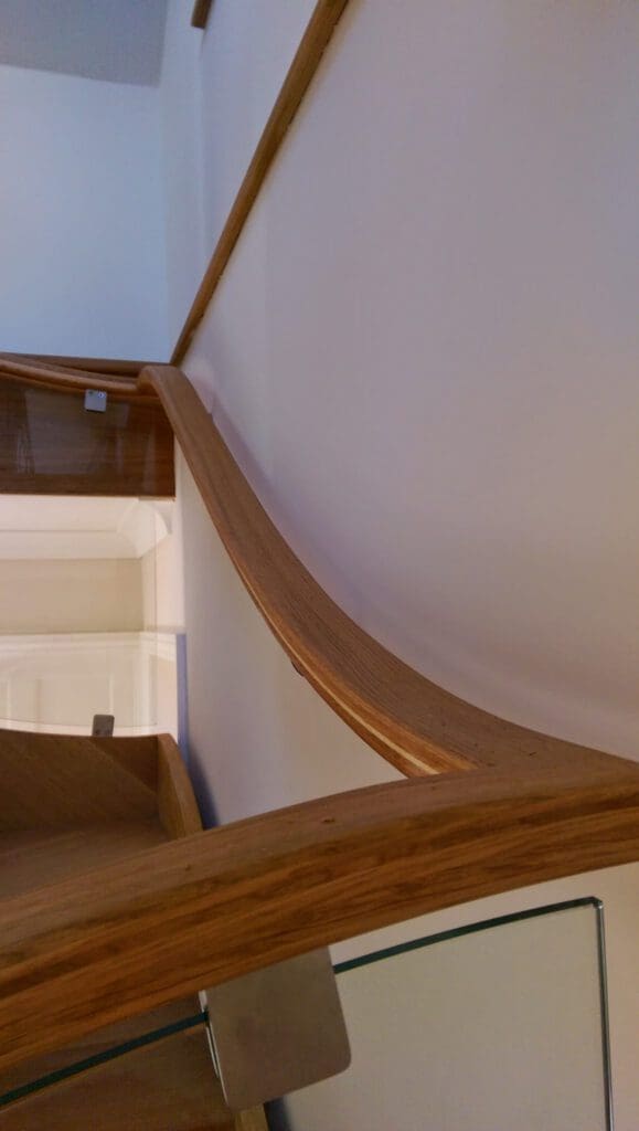 A close-up view of a modern staircase with a smooth, curved wooden handrail, white walls, and glass panels with metal brackets. The photo is taken looking upward along the handrail.