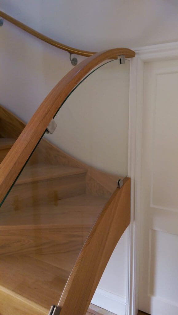 A modern wooden staircase with a curved handrail and glass balustrade, leading upward next to a white wall and door. The design features clean lines and a natural wood finish.