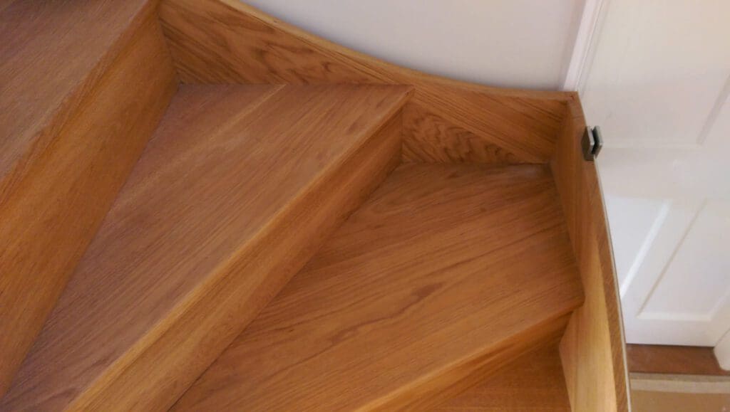 Close-up view of wooden stairs with angular, triangular steps, bordered by wooden trim and a white wall with a white door partially visible in the background.