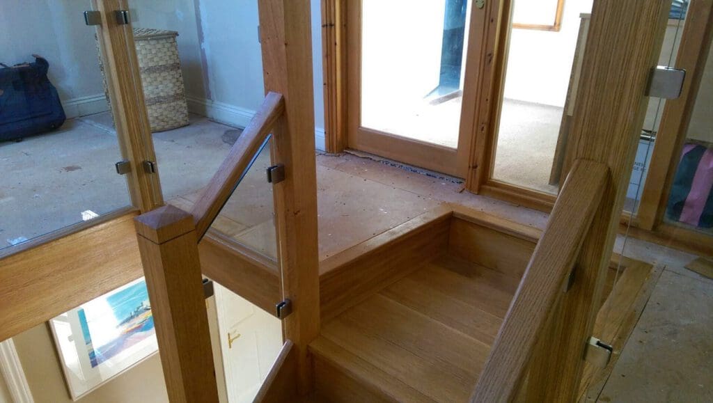 Wooden staircase with glass panel railings leading to a landing area in a house under renovation; visible are unfinished floors, a laundry basket, and some construction materials.