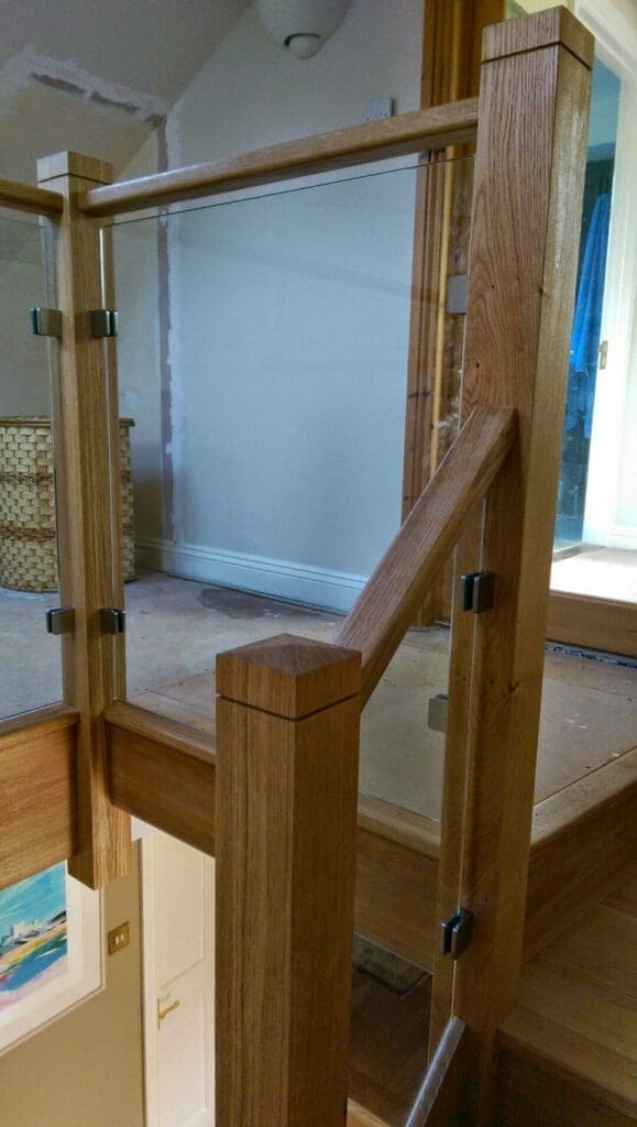 A staircase with a wooden handrail and vertical posts, featuring glass panels held in place by metal brackets. The area is well-lit, and the stairs lead to a hallway with unfinished flooring.