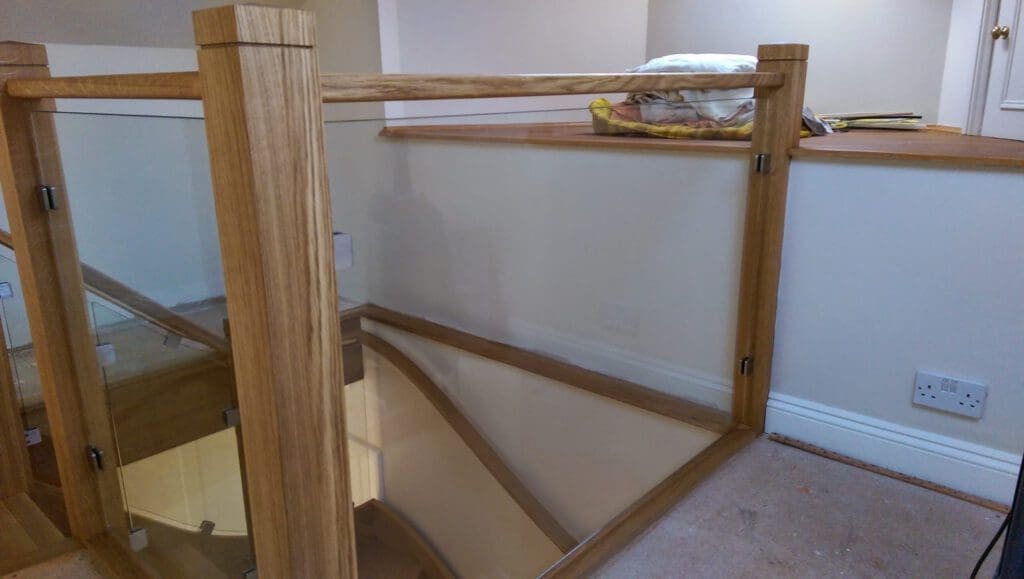 A wooden staircase with glass panel railings, leading downward. The landing area has beige walls, a carpeted floor, a pile of folded linens on a ledge, and a power outlet visible on the wall.